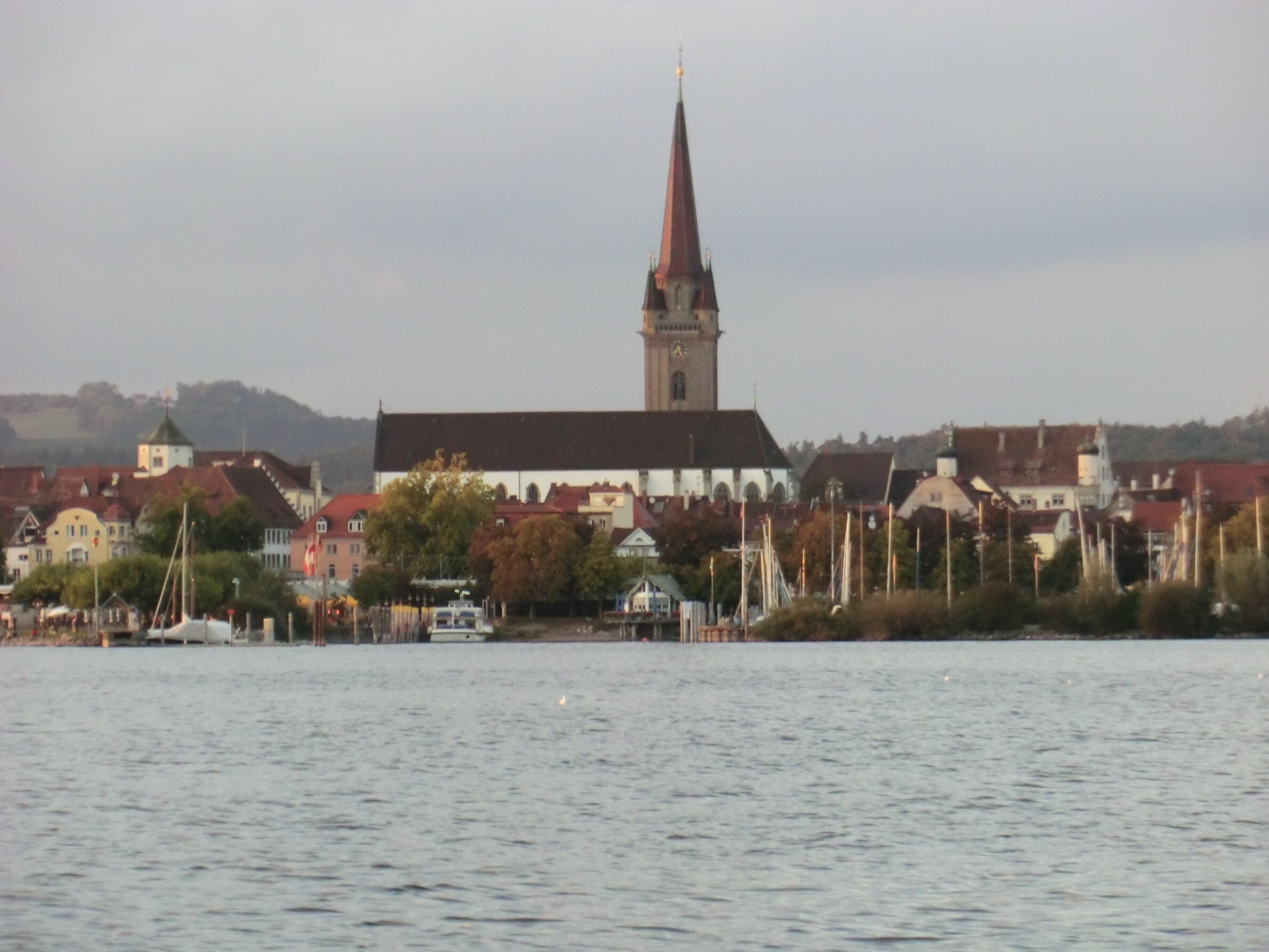 Een kerk met een hoge spits en een klokkentoren, omgeven door gebouwen met rode daken, aan de overkant van een waterpartij.