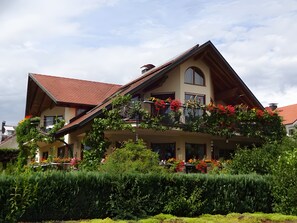 Una casa de dos pisos con techo inclinado, un balcón adornado con flores rojas y un jardín bien cuidado.