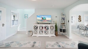 A living room with a TV on a white cabinet, a bookshelf, and a dining area with a table and chairs. - Mariner's Light by Seaside Vacations & Sales (Chincoteague Island)