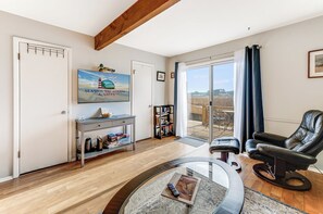 A spacious living room with a large TV, a comfortable leather chair, a glass coffee table, and a bookshelf. - The Teal on Eel Creek by Seaside Vacations (Chincoteague Island)