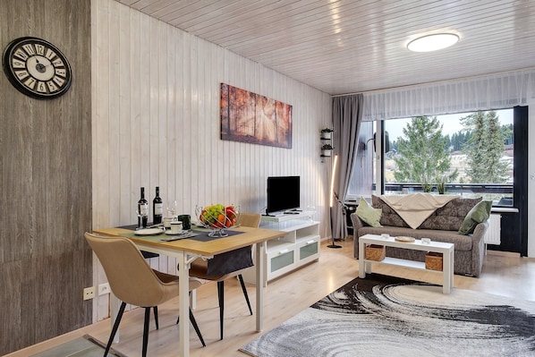 Ein modernes Wohnzimmer mit Essbereich, einem Flachbildfernseher und einem großen Fenster mit Blick auf Bäume.
