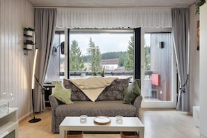 Ein gemütliches Wohnzimmer mit einer Couch, einem Couchtisch und Blick auf Bäume und ein Gebäude durch ein großes Fenster.