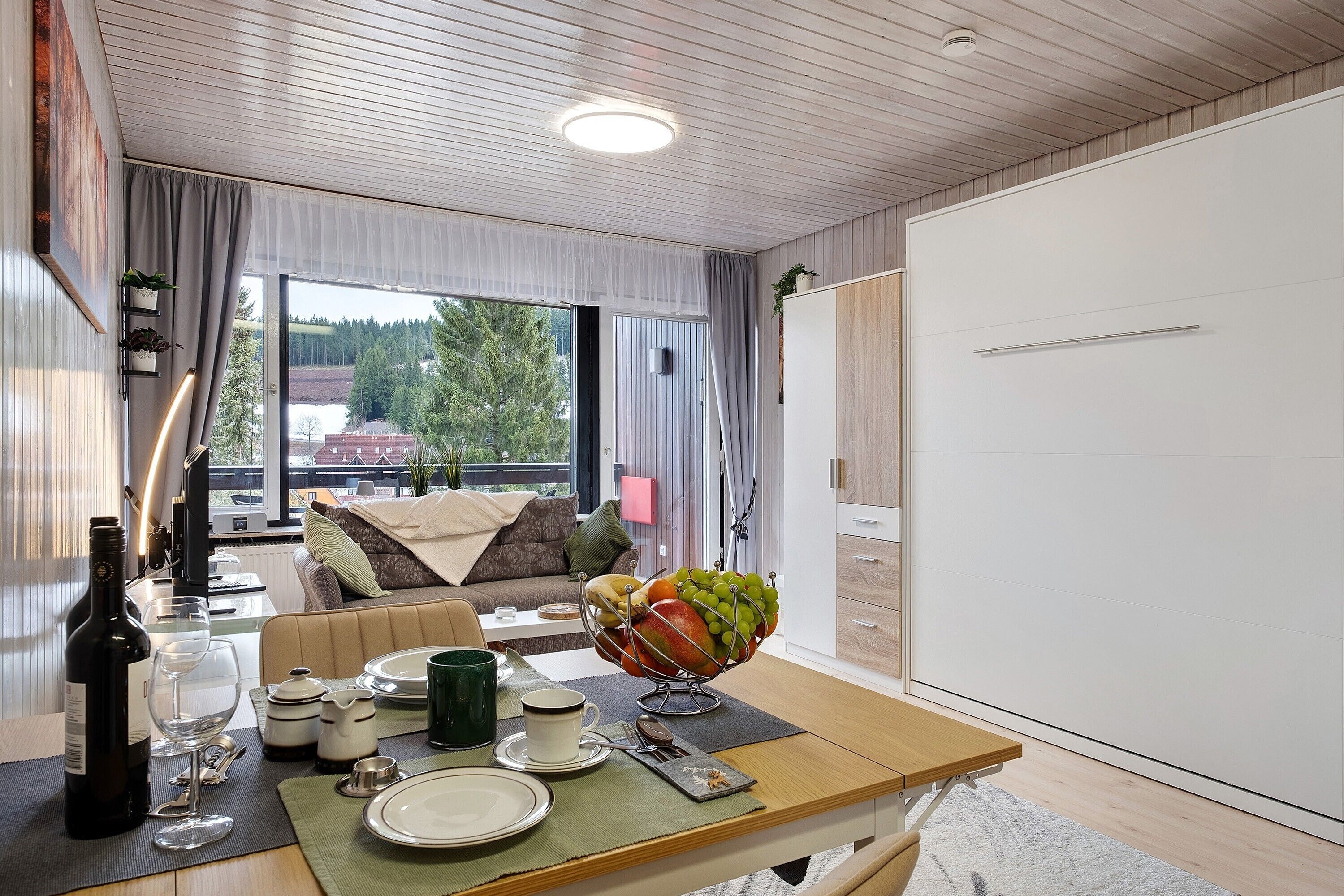 Ein modernes Zimmer mit Esstisch für zwei Personen, einer Couch, einem großen Fenster mit Blick auf Bäume und einem weißen Kleiderschrank.
