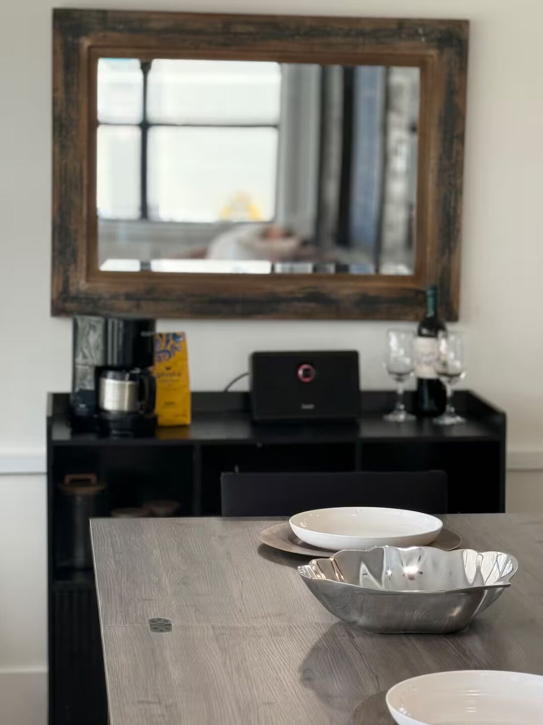 A dining table with a modern design, a large mirror above it, and a black console table with a coffee maker, wine glasses, and a bottle.