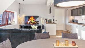 A modern living room with a flat-screen TV, a sofa, a dining table with candles, and a kitchen in the background. - The Summit Rooftop Spa Meissen – Exclusive Penthouse with Jacuzzi & Sauna (Meißen)