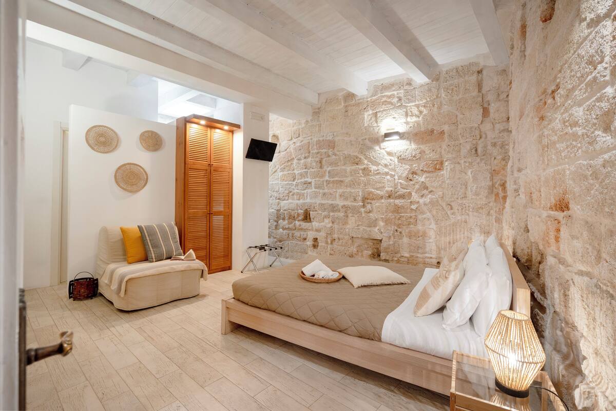 Une chambre avec un mur en pierre, un lit avec une literie blanche, un banc en bois et une petite table avec une lampe.