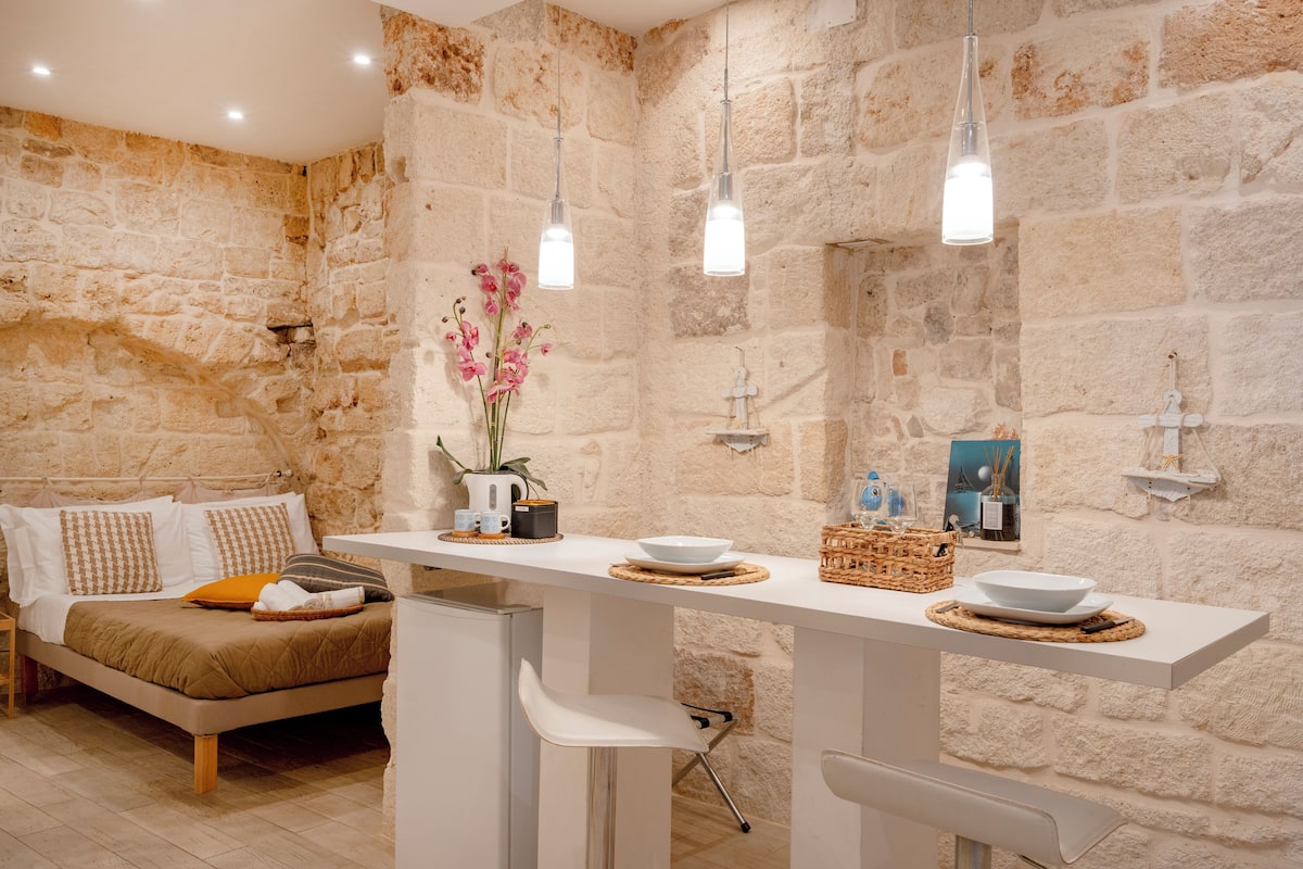 Une chambre moderne avec un mur en pierre, un lit avec une literie blanche et marron, une table blanche avec un bol et un vase de fleurs, et des luminaires suspendus.