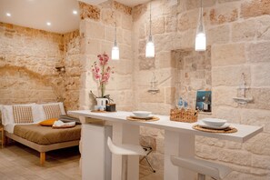 Une chambre moderne avec un mur en pierre, un lit avec une literie blanche et marron, une table blanche avec un bol et un vase de fleurs, et des luminaires suspendus.