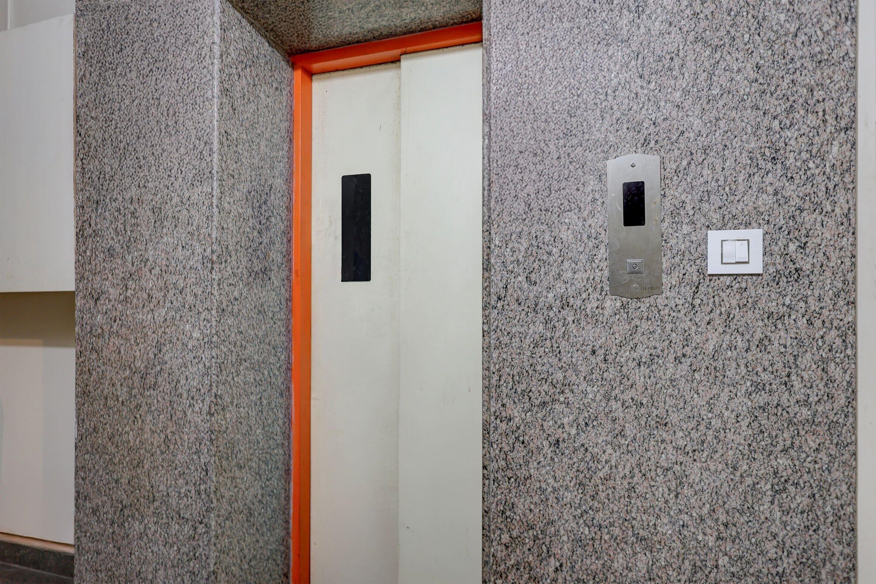 Un ascenseur avec un mur en granit, une porte ornée d’un rectangle noir et un interrupteur.
