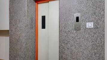 Un ascenseur avec un mur en granit, une porte ornée d’un rectangle noir et un interrupteur.