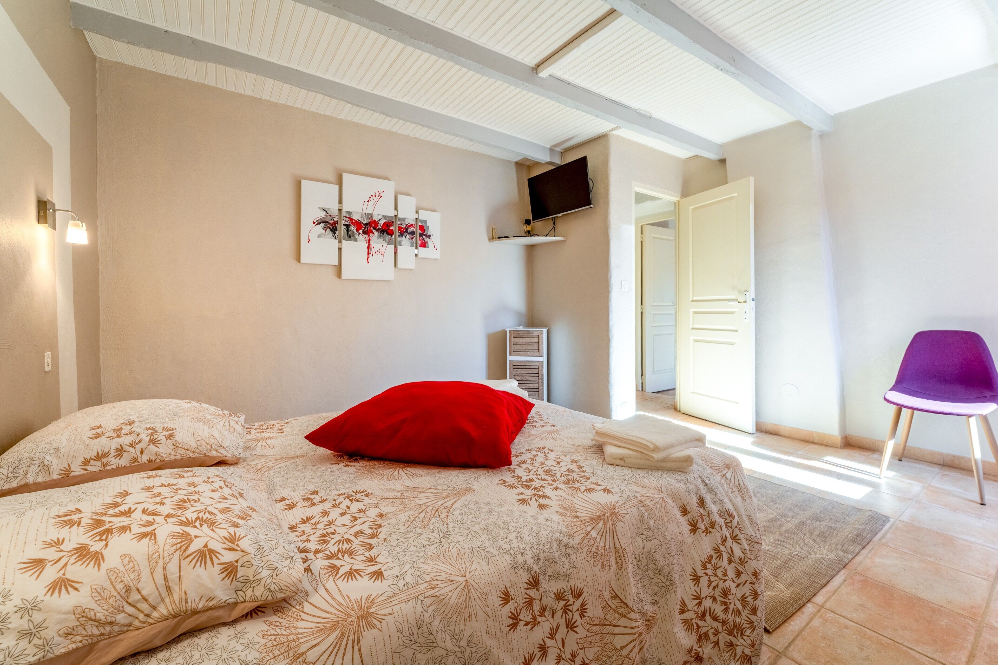 Un dormitorio con una cama, una almohada roja, un televisor, una silla y una puerta que da a otra habitación.