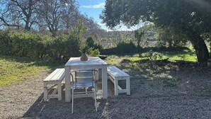 A wooden table and two chairs set on a gravel surface, surrounded by trees and greenery.