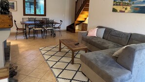 A living room with a grey sofa, a wooden coffee table, and a dining area with a wooden table and chairs. There is a staircase leading to an upper level, and a painting of a beach scene on the wall.