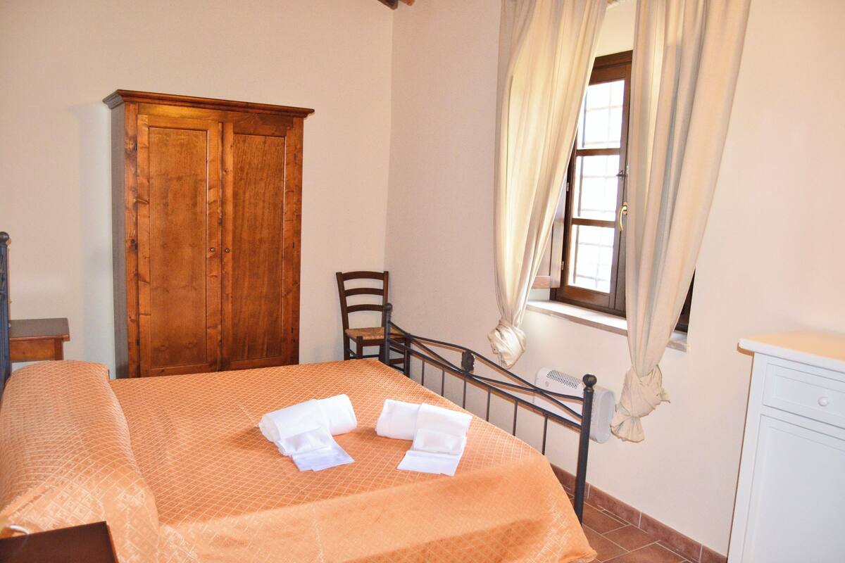 Un dormitorio con cama, armario de madera, una silla y ventana con cortinas.