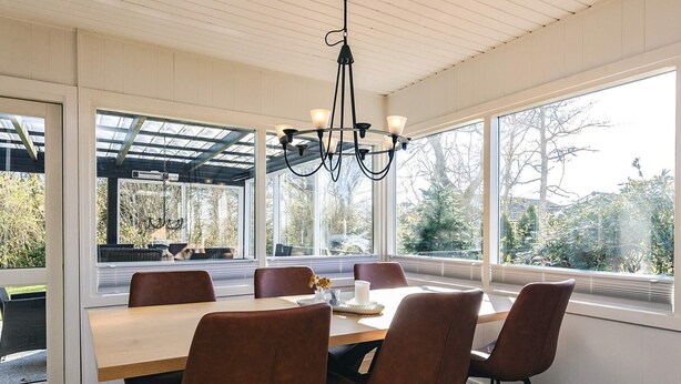 A dining area with a wooden table, six chairs, a chandelier, and large windows with a view of trees. - 6 Person Holiday Home in Logstor (Logstor)
