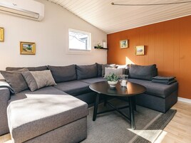 Sala de estar moderna com um sofá seccional cinza, uma mesa de centro redonda e uma pequena mesa lateral. Há uma janela com vista para o exterior, e a parede apresenta painéis de madeira.
