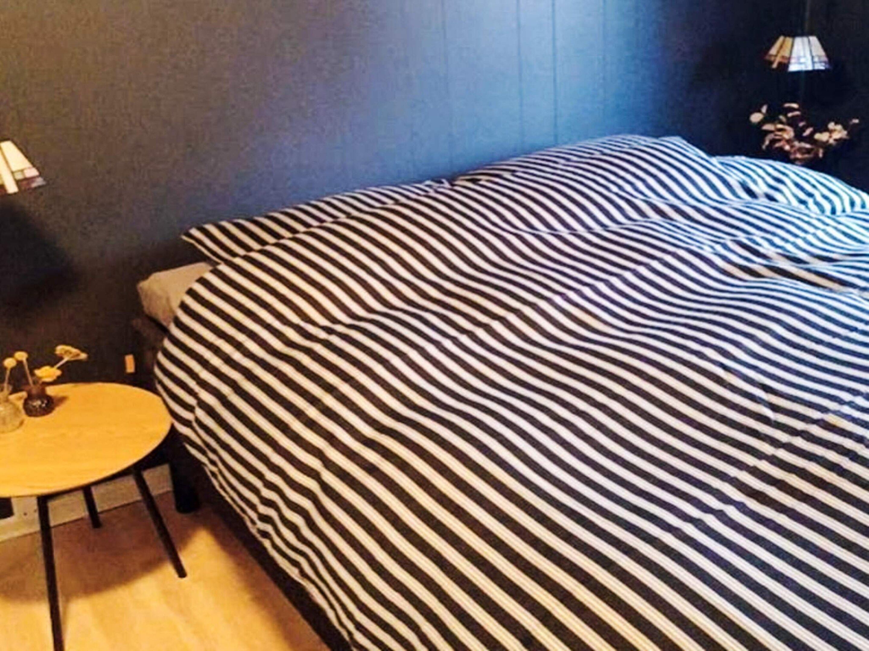 A room with a bed covered in striped bedding, a small round table with a vase, and a lamp on a nightstand.