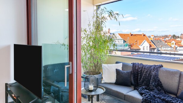 A balcony with a sofa, a TV, a potted plant, and a view of buildings. - The Summit Suite I Meissen – Luxury Penthouse with Elbe and Cathedral Views (Meißen)
