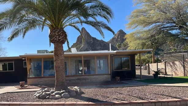 Una casa moderna con ventanas amplias, una palmera y montañas rocosas al fondo.