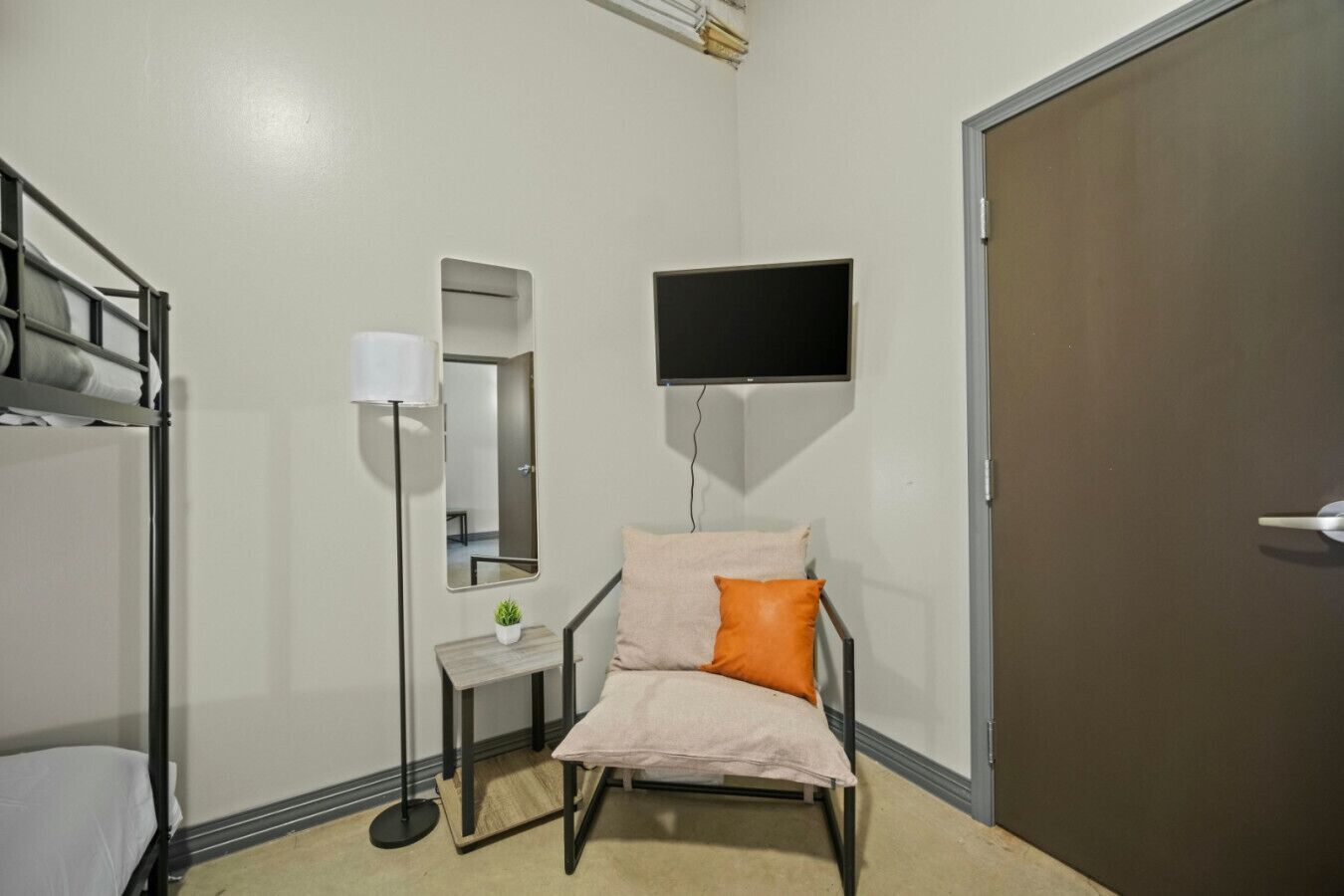 A compact room with a bunk bed, a desk with a lamp, a chair with cushions, a flat-screen TV, and a door.