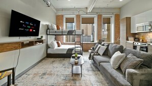 Un espacio habitable compacto con una cama nido, un sofá, una mesa de centro, un televisor montado en la pared y una zona de cocina.
