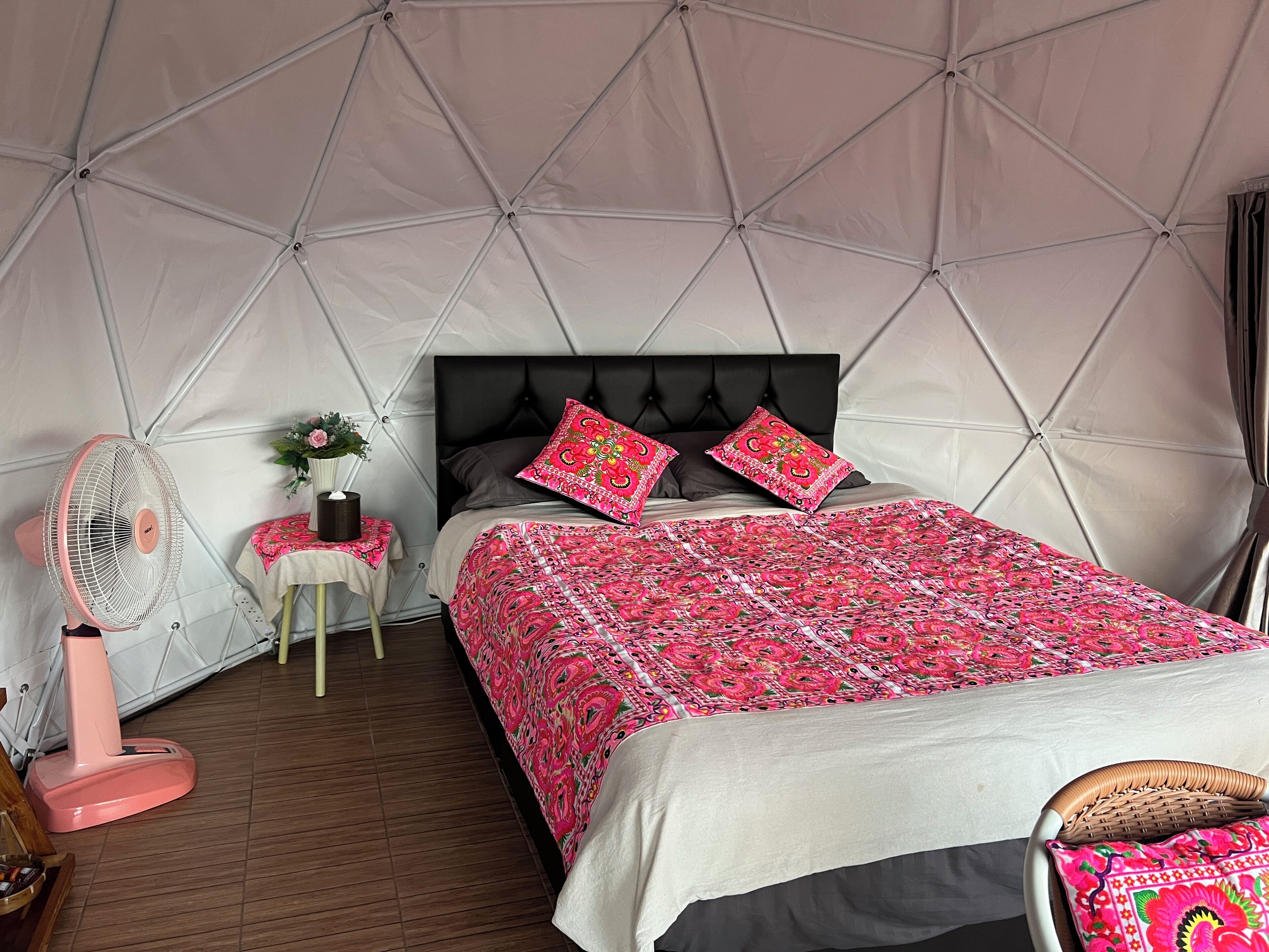 A tented room with a bed, a fan, a small table with a vase, and a chair.
