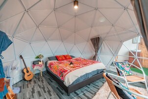 Una habitación con carpa, una cama, una guitarra, un ventilador y una pequeña mesa con un radio.