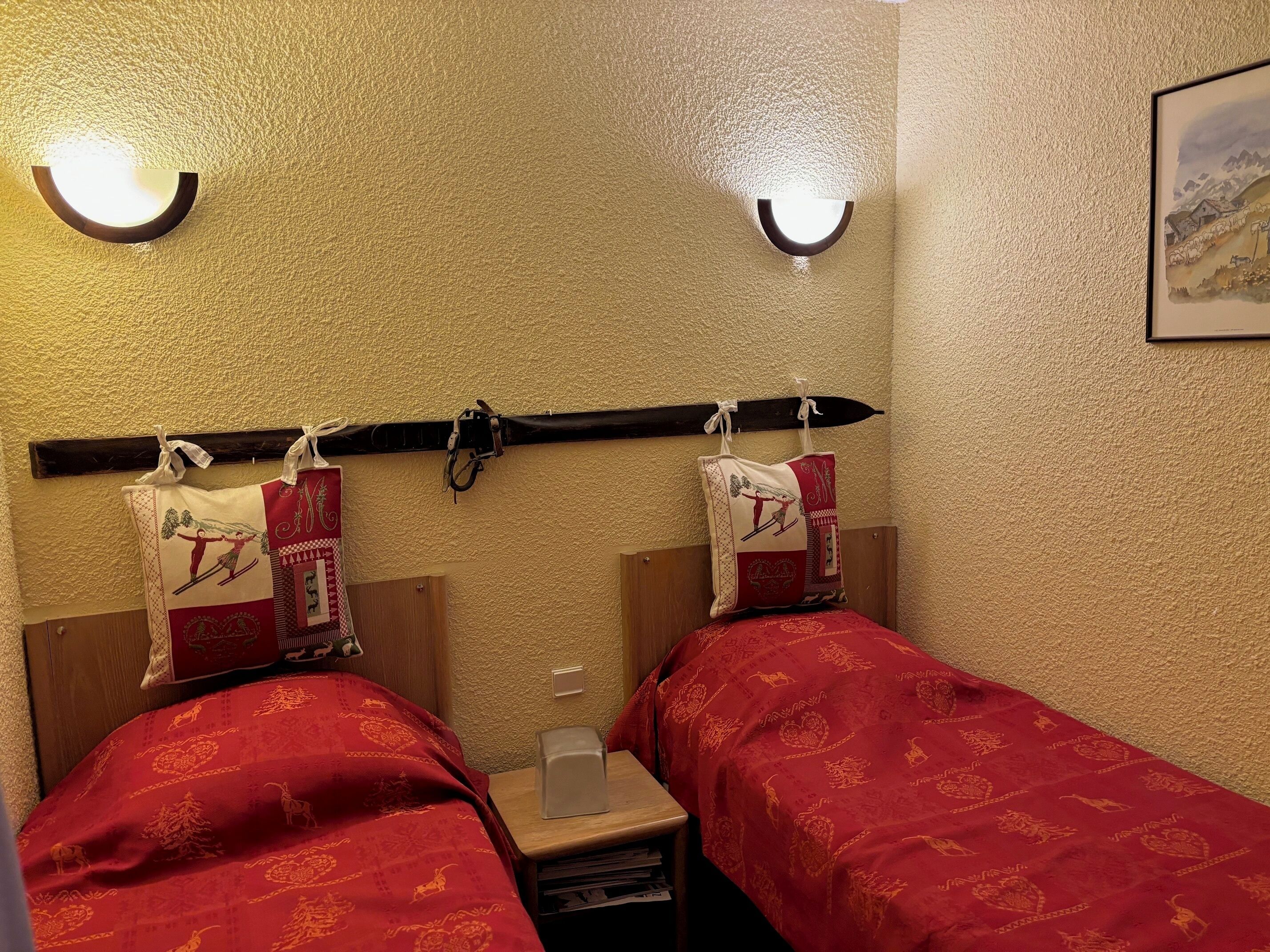 A hotel room with two beds, each with a red bedspread and matching pillows. There are wall-mounted lights above the beds and a painting on the wall.