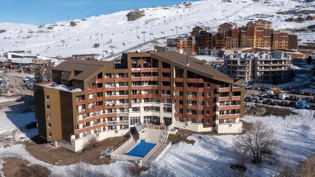 Een meerhoog hotel met een zwembad op de voorgrond, omringd door met sneeuw bedekte bergen en skipistes.