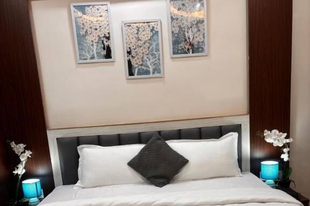 Una cama bien hecha con ropa de cama blanca y gris, un cabecero oscuro y tres cuadros enmarcados encima.