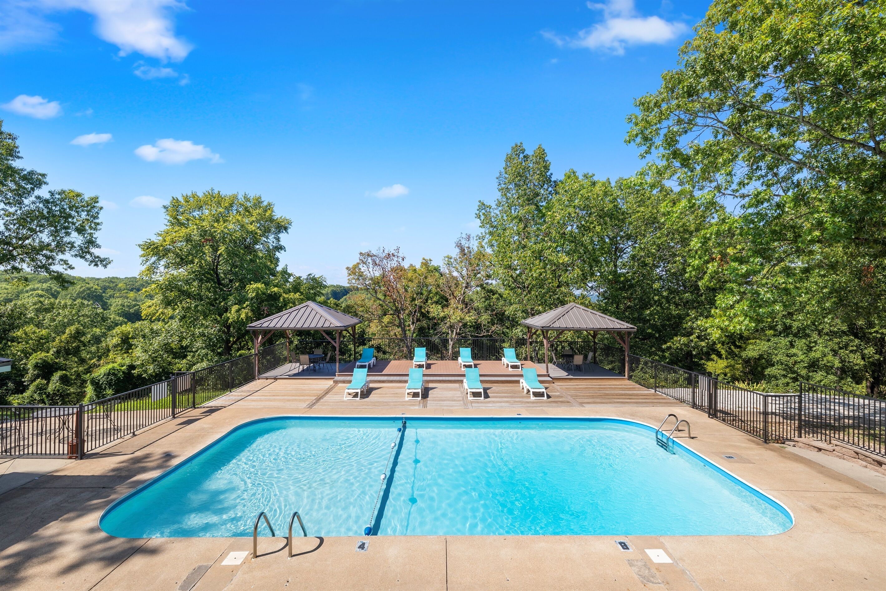 A rectangular swimming pool with a diving board, surrounded by lounge chairs and shaded areas, set against a backdrop of trees and a clear sky.