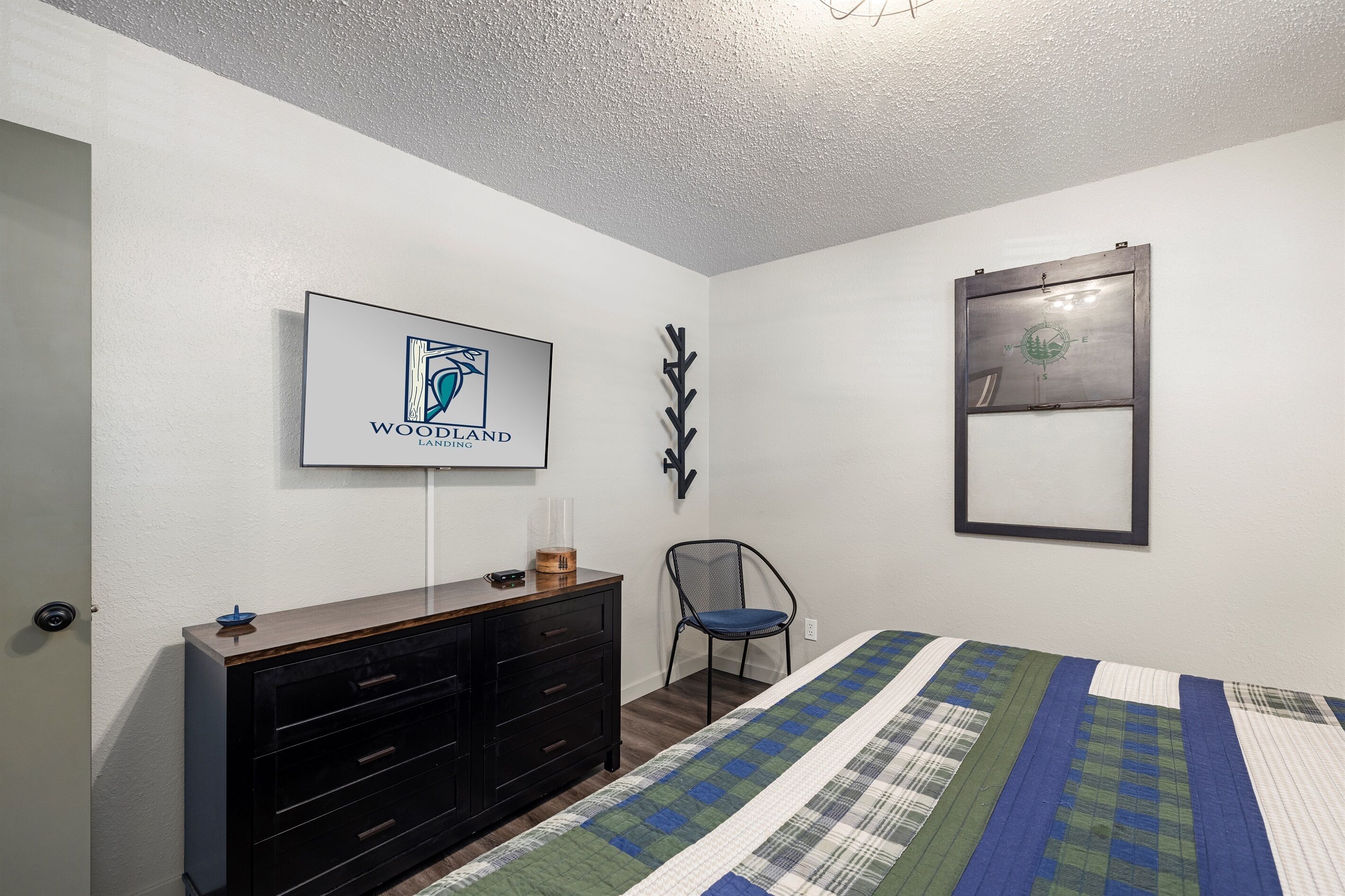 Une chambre à coucher comprenant un lit, une commode, une chaise, un téléviseur fixé au mur et un tableau encadré.