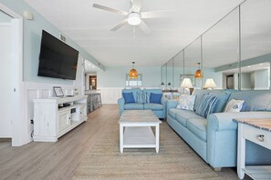 A spacious living room with a large light blue sofa, a white coffee table, and a ceiling fan. - Oceanfront condo sleeps 8, open for summer! (Panama City Beach)