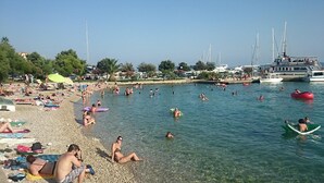 Een druk strand met mensen die zwemmen en zonnebaden, boten afgemeerd in de buurt, en een heldere lucht.