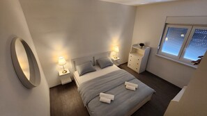 A modern bedroom with a large bed, bedside tables with lamps, a dresser, and a window with blinds. - HIDDEN SUNSET - one bedroom sea front island aparment (Selca)