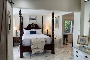 Una cama con dosel, con cubrecama blanco y almohada azul, una mesita de noche con lámpara, y un cuadro enmarcado en la pared.