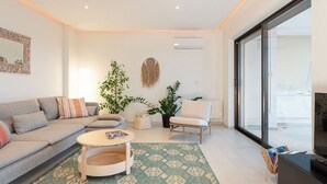 Un salon moderne avec un canapé, une table basse ronde, un fauteuil et un téléviseur. On y trouve également des plantes en pot et une grande fenêtre.