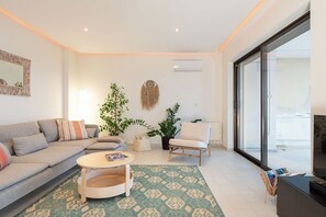 A modern living room with a sofa, a round coffee table, a chair, and a TV. There are potted plants and a large window. - South Blue Seafront Vouliagmeni spacious Apartment (Vouliagmeni)