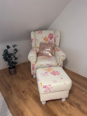 Un sillón estampado con motivos florales, un reposapiés a juego, una planta en maceta metálica y suelo de madera.