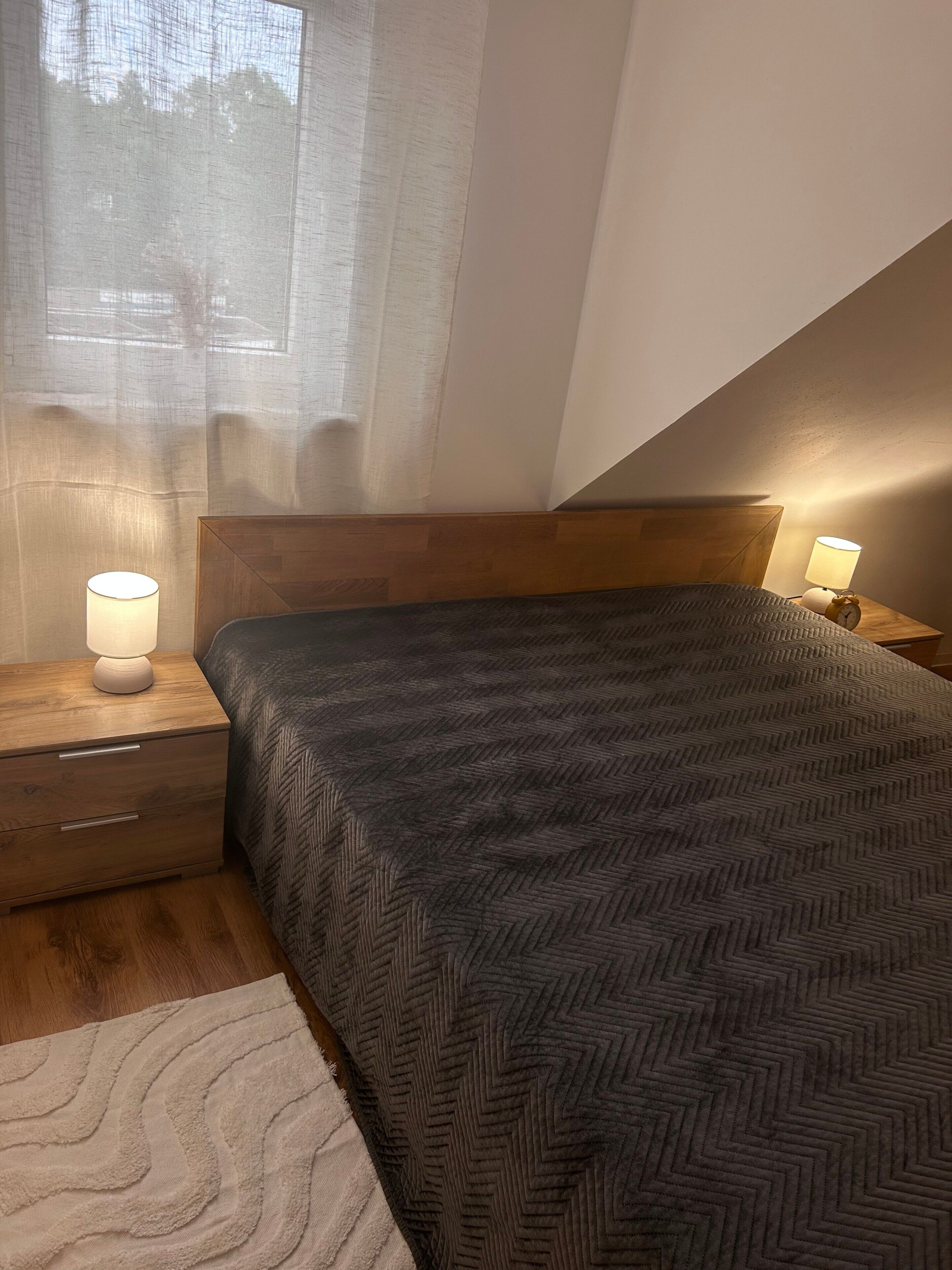 A bedroom with a bed, two bedside lamps, a wooden headboard, and a window with sheer curtains.