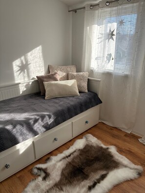 Un dormitorio con cama, almohadas, un manto de piel sintética, una ventana con cortinas y un radiador.