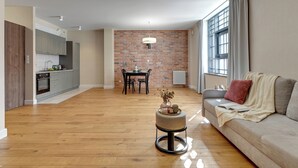 A modern living room with a wooden floor, a grey sofa with a red pillow, a round ottoman with a vase, and a brick wall behind a dining table with chairs. - Maszynownia by BrowarHevelius Apartments (Gdańsk)