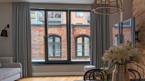 A modern living room with a large window, a sofa, a dining table, and a vase of flowers. - Maszynownia by BrowarHevelius Apartments (Gdańsk)