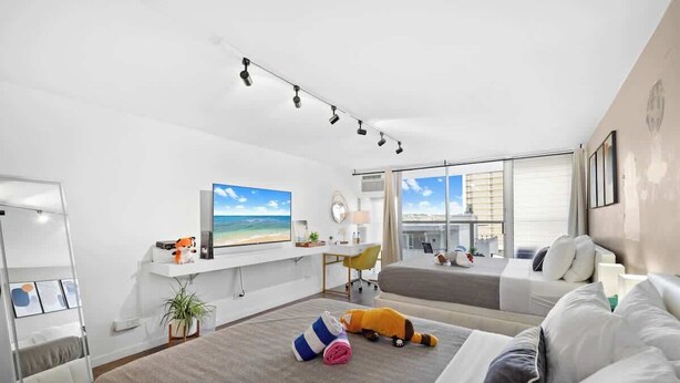 A modern hotel room with a large bed, a desk with a computer, a TV, and a view of the city. - @ Marbella Lane Chic Seaside Studio w/ Pool View (Honolulu)