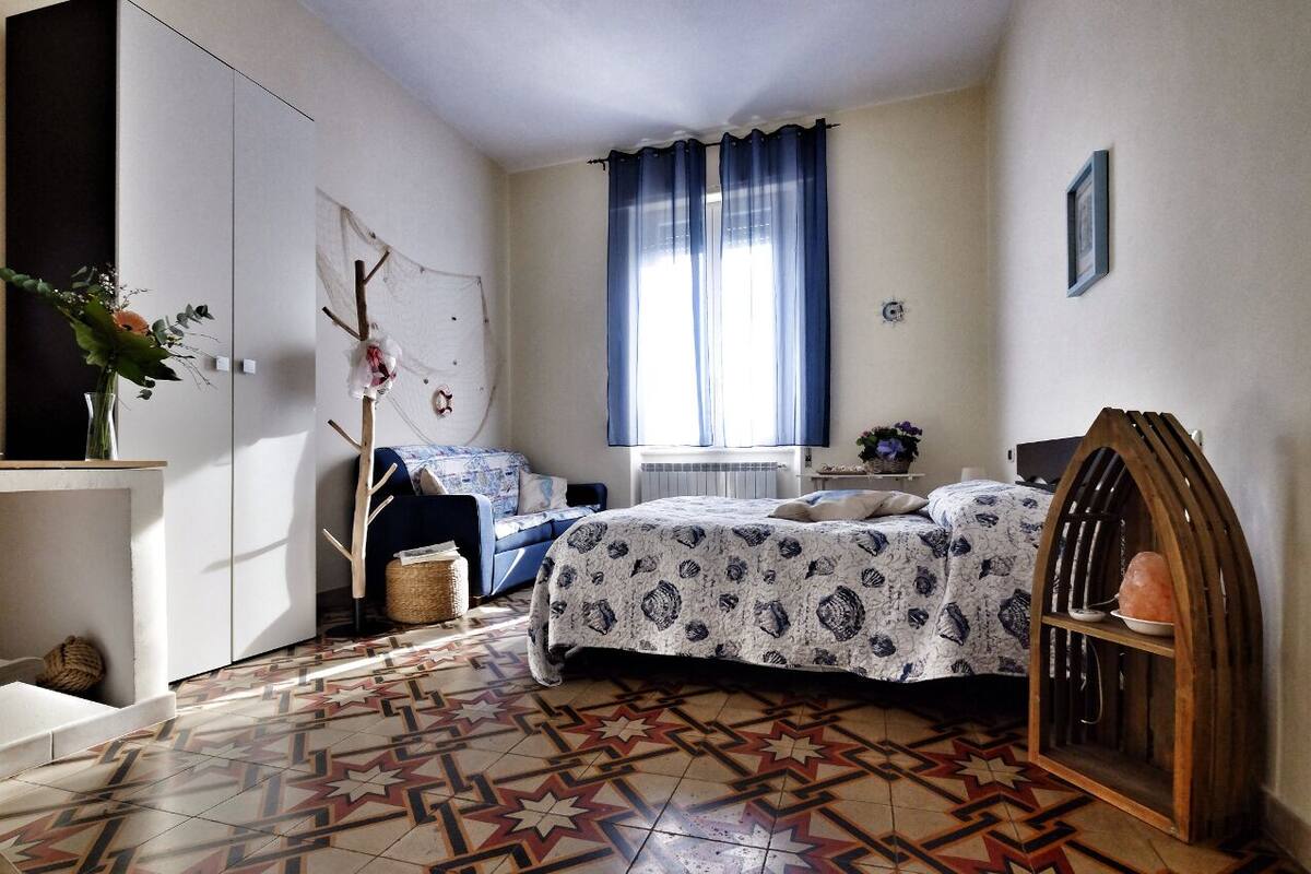A bedroom with a patterned floor, a bed with a patterned bedspread, a window with blue curtains, a bedside table with a lamp, and a wall-mounted shelf.