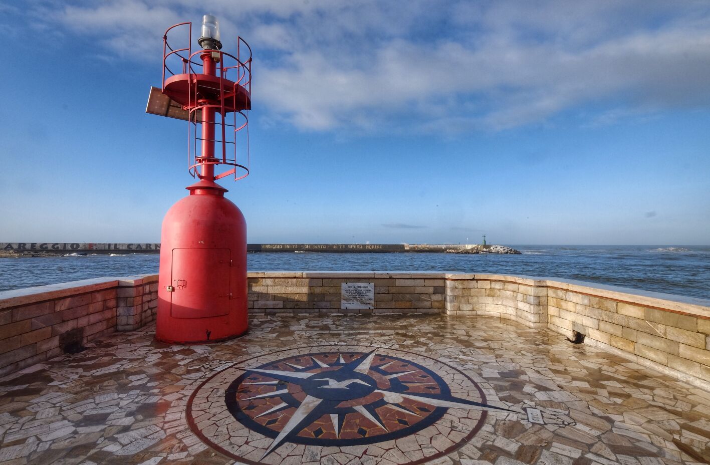 A red lighthouse on a tiled platform with a compass mosaic, overlooking the sea.