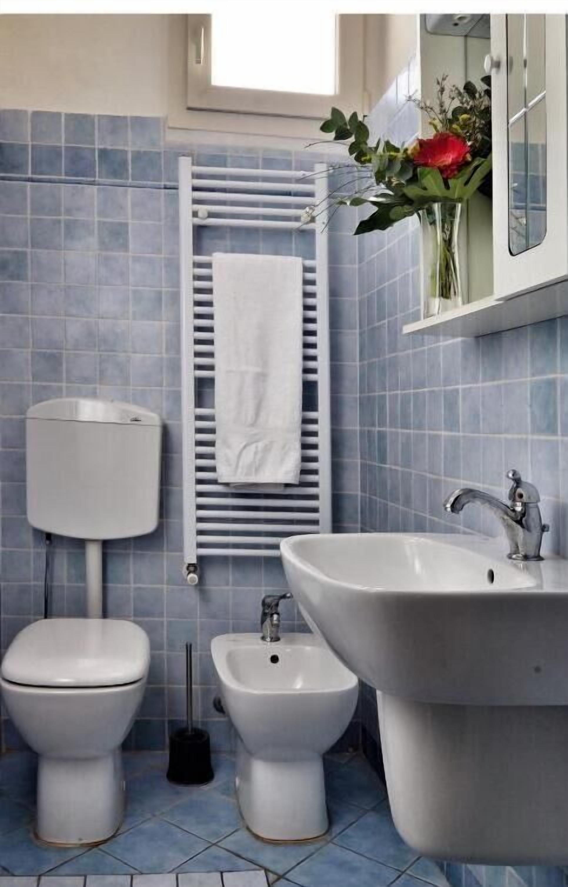 Casa de banho com sanita, bidé e lavatório, revestida a azulejos azuis, com um porta-toalhas e um vaso de flores.