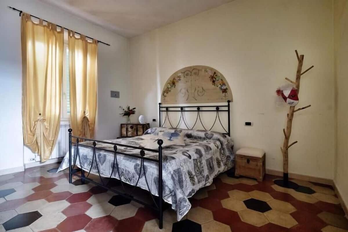 A bedroom with a bed, curtains, a decorative wall piece, a small wooden chest, and a unique floor pattern.