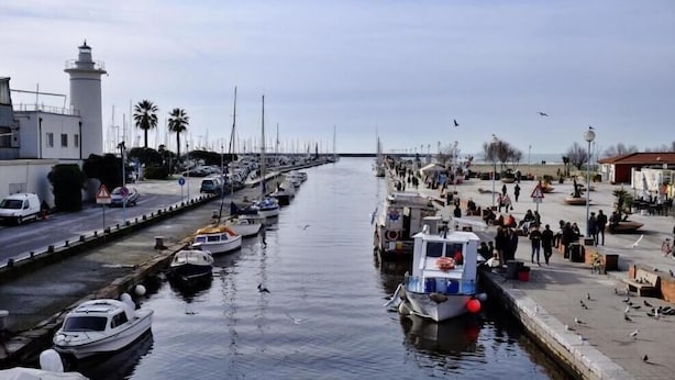 Una marina con barcos amarrados a lo largo del agua, un faro al fondo y un paseo marítimo con gente caminando.