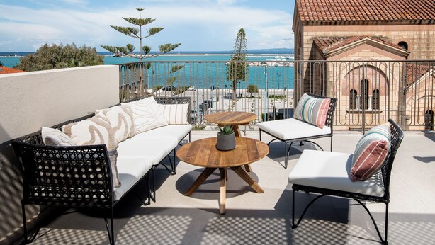 Eine Terrasse mit einer Couch, Sesseln und einem Tisch, mit Blick auf einen Strand und ein Gebäude mit einem Ziegeldach.